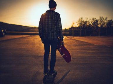 Junger Mann mit Skateboard bei Sonnenuntergang, blickt in die Ferne.